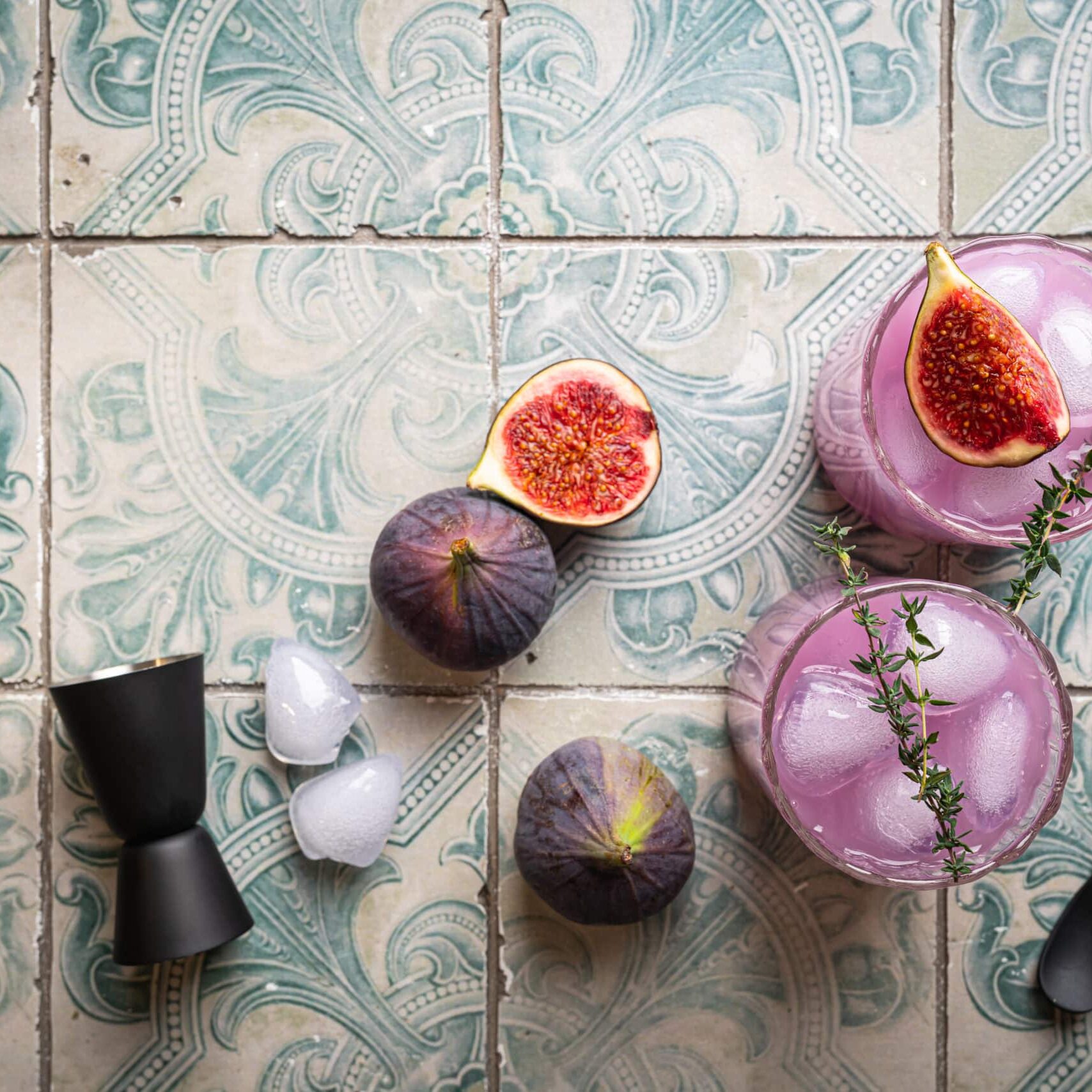 Purple fig cocktail with ice and thyme on tile background, top view